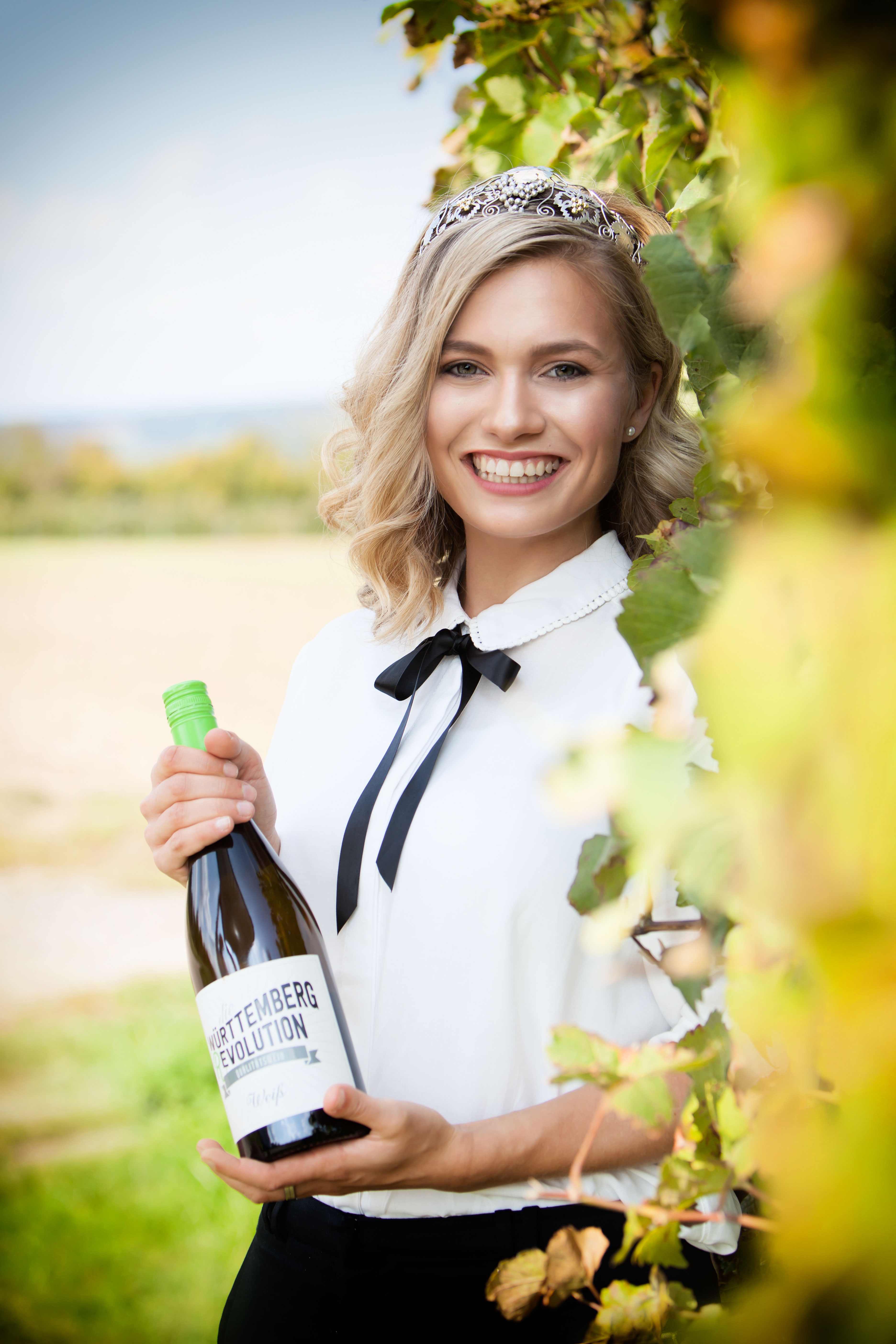 Man sieht die Württemberger Weinkönigin Carolin Klöckner, mit einer Weinflasche, vor einem Feld.
