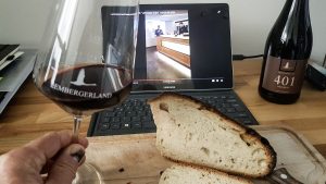 Live Tastings bei der Lembergerland Kellerei. Man sieht einen aufgeklappten Laptop, vor dem ein geschnittenes Brot liegt. Eine Person hält ein Glas Rotwein in der Hand.