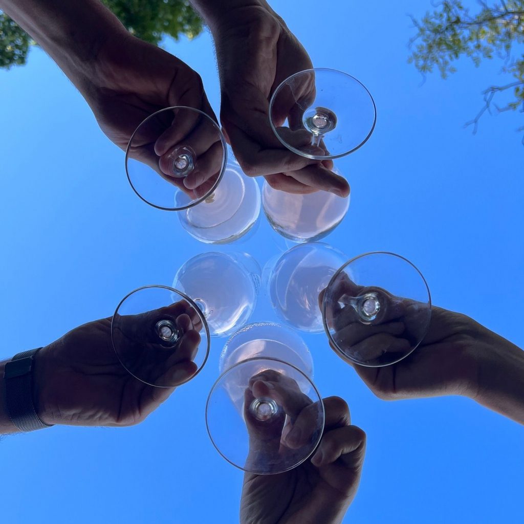 Mehrere Menschen, die man im Bild aber nicht sieht, stoßen mit Weingläsern an. Von unten fotografiert, im Hintergrund der blaue Himmel.
