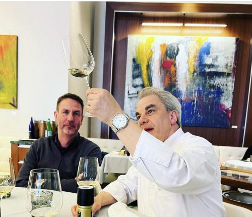 Dr. Kost mit Gabriel Kreuther am Tisch von dessen Restaurant, Kreuther hält Glas in die Luft und schaut prüfend den Pinot Meunier Sparkling Rosé an