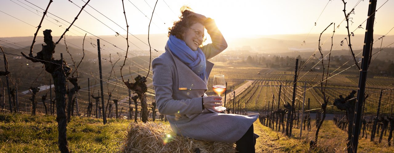 Anja Off sitzt auf einem Strohballen im Weinberg, sie hat eine Flasche Orange Wine dabei und ein Glas davon in der Hand, im Hintergrund geht die Sonne unter