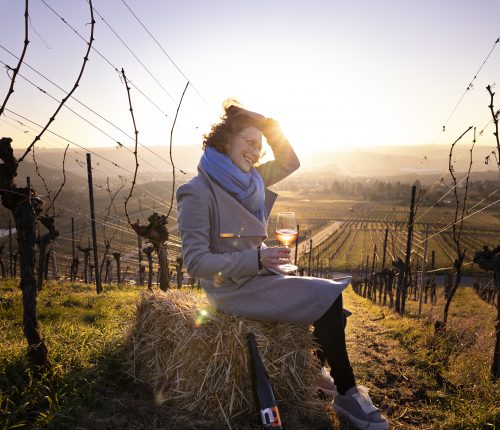 Anja Off sitzt auf einem Strohballen im Weinberg, sie hat eine Flasche Orange Wine dabei und ein Glas davon in der Hand, im Hintergrund geht die Sonne unter