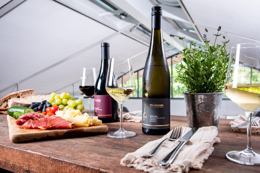 Auf einem Holztisch steht eine Vesperplatte mit Schinken, Käse, Trauben, Tomaten und Gurke, dazu Brot und zwei Flaschen Wein der Fellbacher Weingärtner aus dem Remstal.