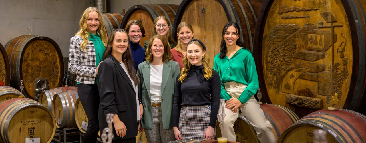 Die fünf Bewerberinnen um das Amt der Württemberger Weinkönigin 2023/24 sitzen zusammen mit den noch amtierenden Weinhoheiten im Weinkeller beieinander.