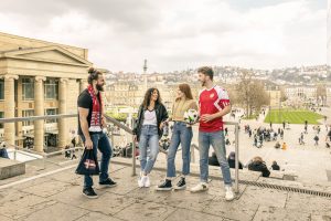 Mehrere junge Menschen stehen oberhalb der Königstraße und des Schlossplatzes in Stuttgart und unterhalten sich, einer trägt ein Fantrikot